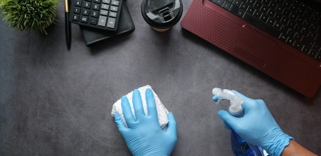 Spring cleaning technology- gloved hands wiping down a desk with a red laptop, calculator and coffee cup on top of desk